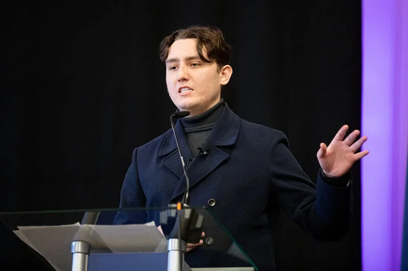 Joseph Roberts speaking and gesturing behind a clear lectern