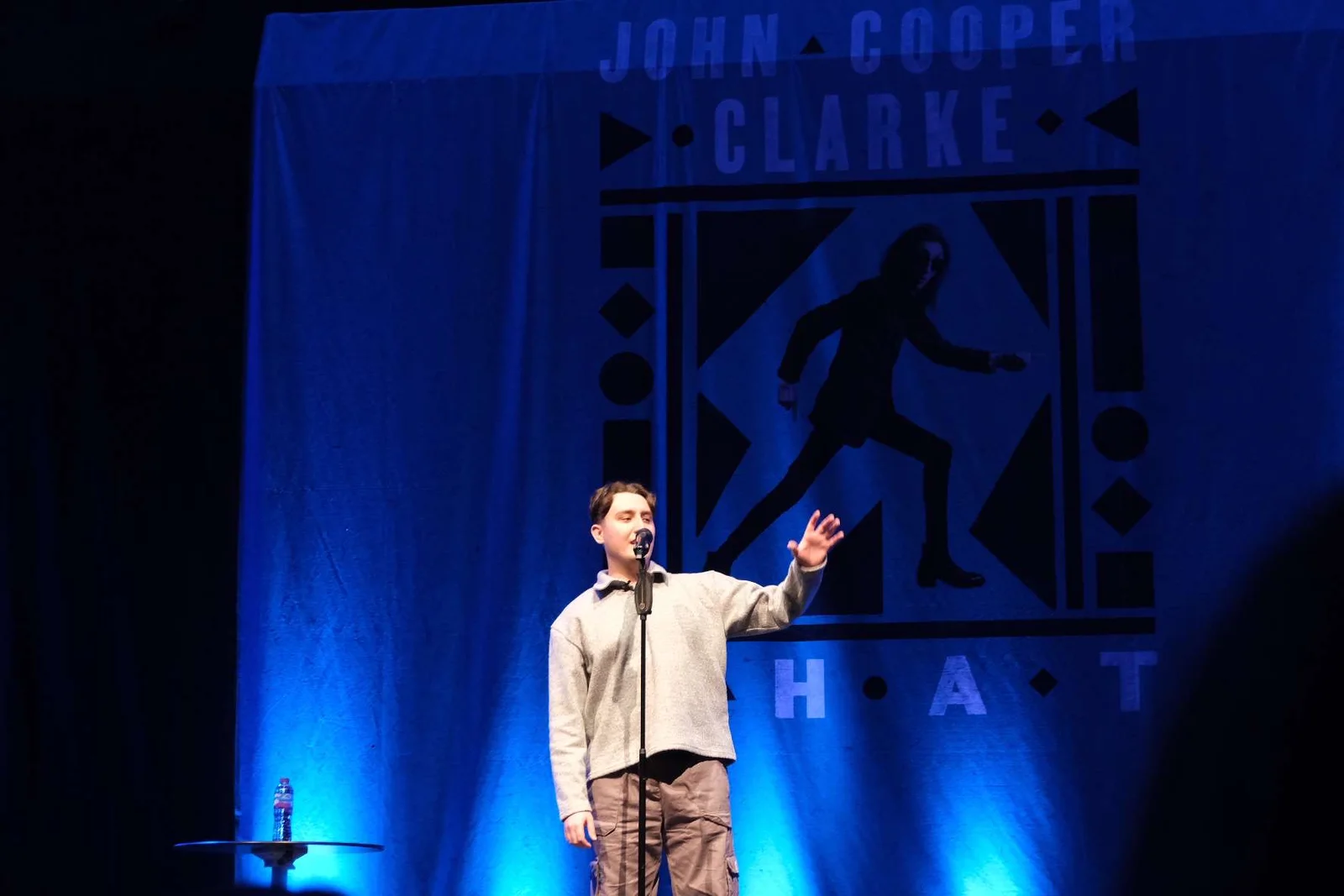 Joseph Roberts performing on a blue-lit stage beneath a John Cooper Clarke backdrop